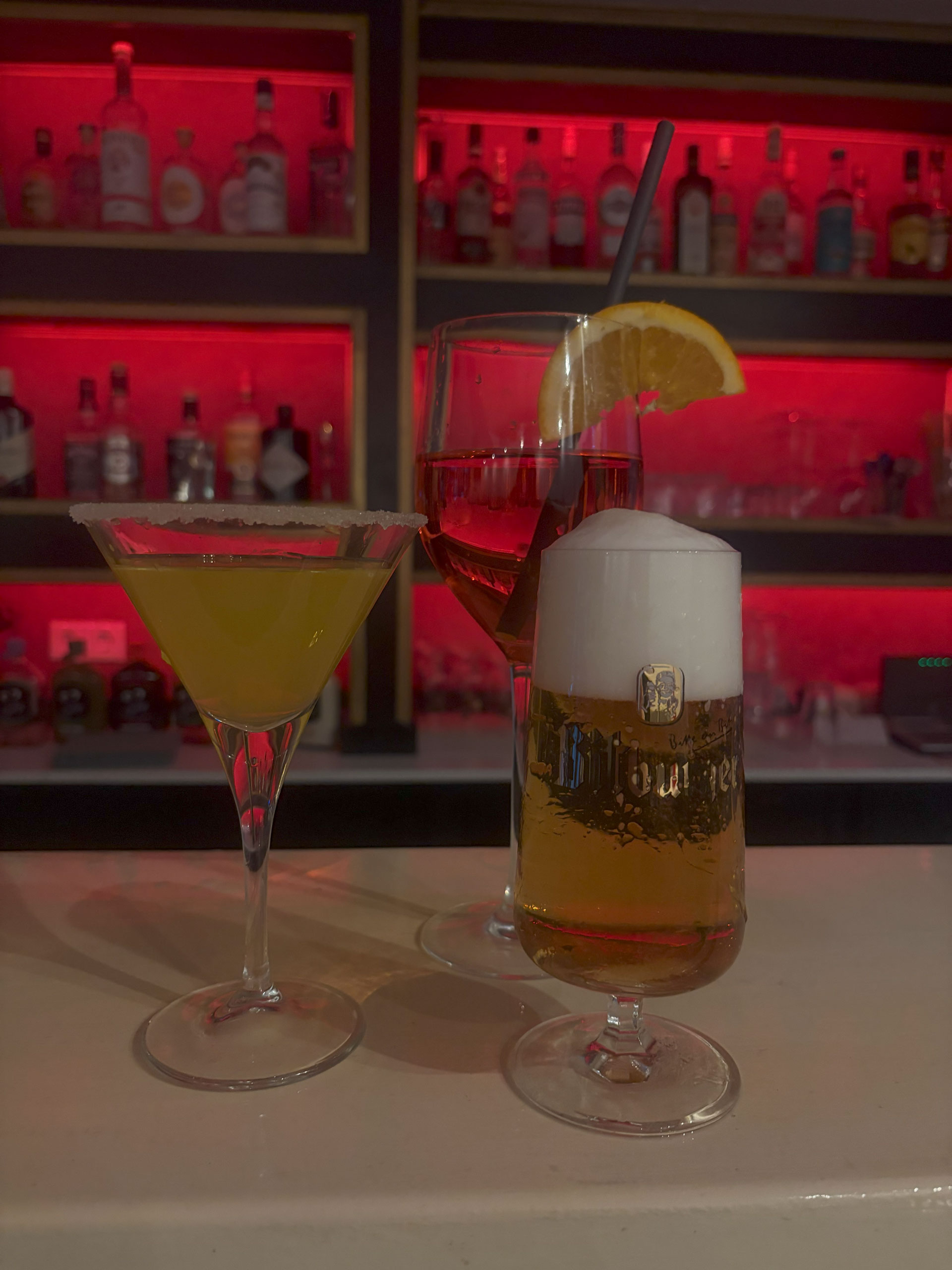 Drei Getränke auf einer Bar, darunter ein Cocktail, ein Bier und ein Wein.